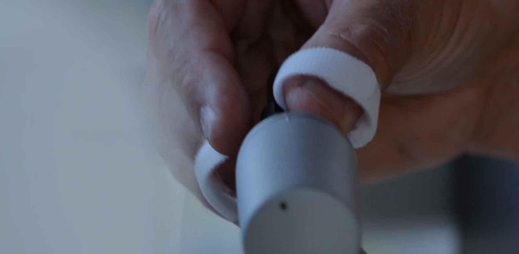 Close-up of a hand with a white wrap around a finger, handling a small gray medical device near the ear.