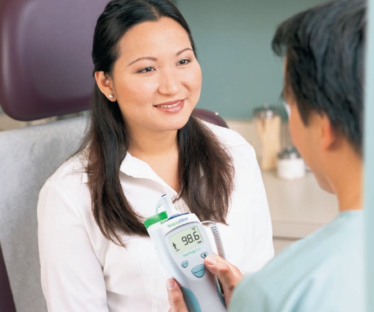 Smiling woman in a white blouse sits in a clinical chair as a clinician uses a digital thermometer showing 98.6°F.