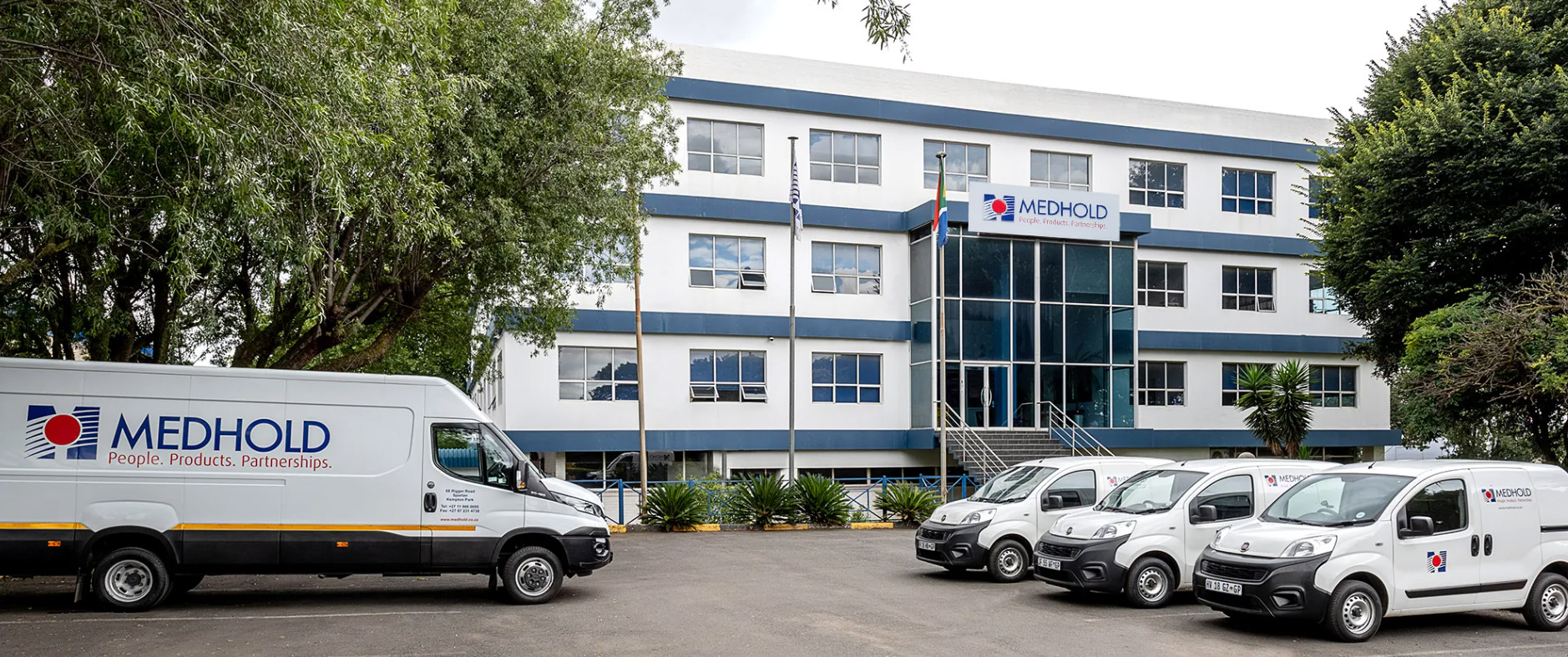 Office building with Medhold sign, glass entrance, and several white company vans parked in front, flanked by trees and flags.
