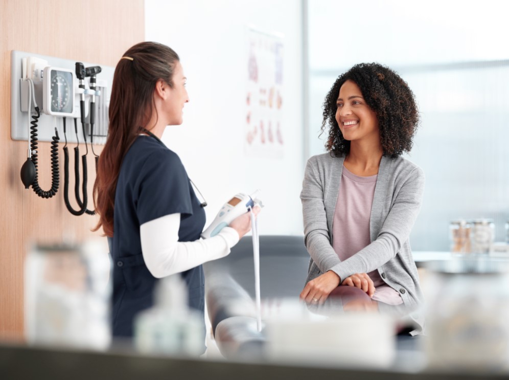 Nurse or clinician speaks with a smiling patient in a clinic exam room, medical equipment on the wall behind them.