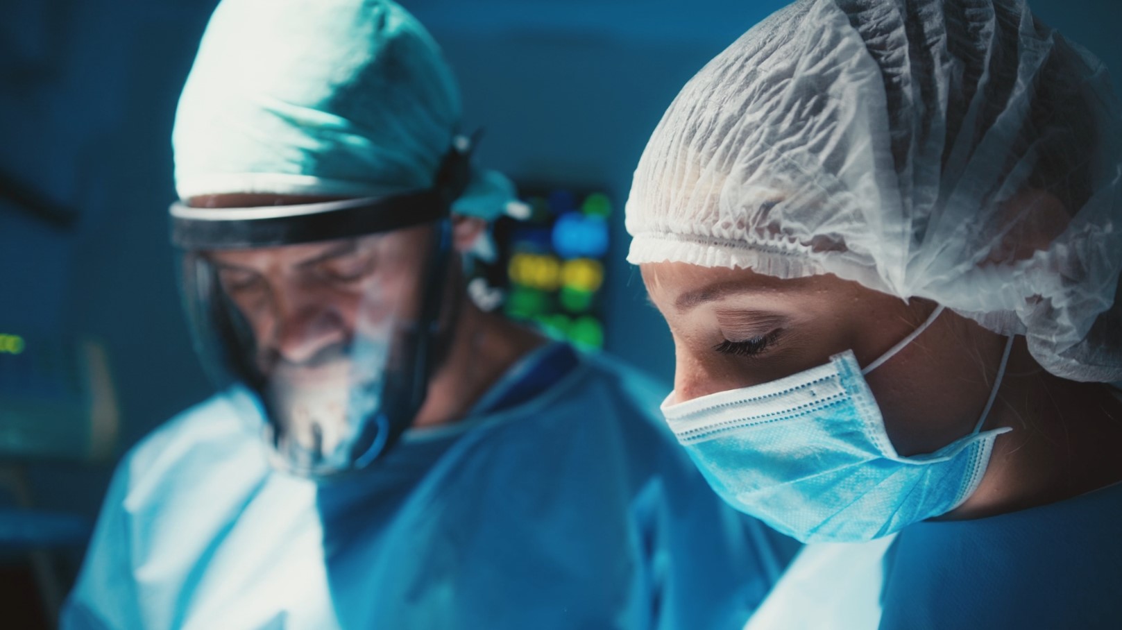 smoke1 Close-up of a female surgeon wearing a blue surgical mask and cap, focused on a procedure with a blurred colleague in the background in an operating room.