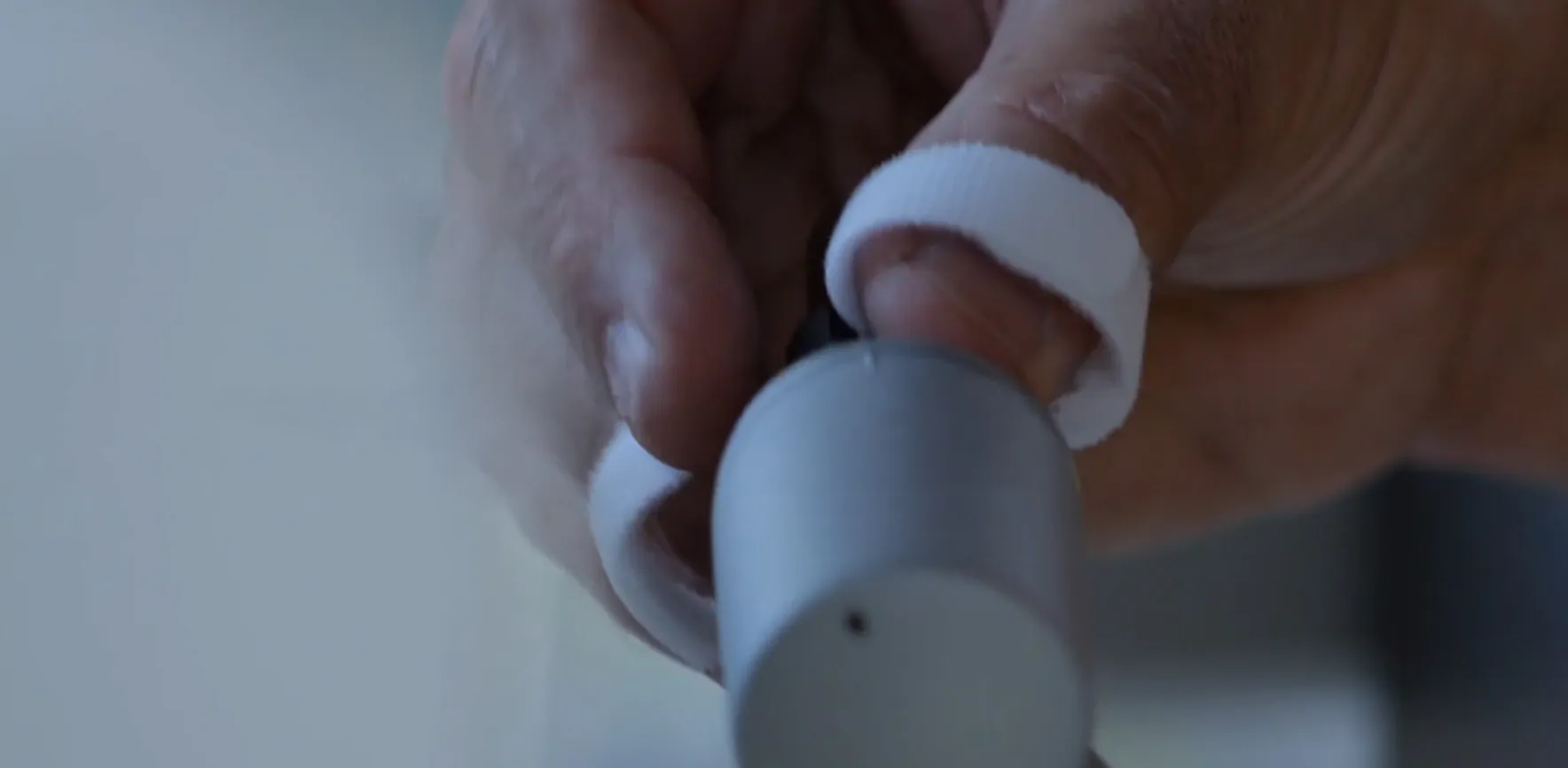 Close-up of a hand with a white wrap around a finger, handling a small gray medical device near the ear.