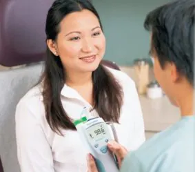 Smiling woman in a white blouse sits in a clinical chair as a clinician uses a digital thermometer showing 98.6°F.