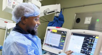 Healthcare worker in blue scrubs and hairnet operating patient monitors in a hospital ICU.