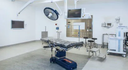 Modern operating room with an empty surgical table, overhead light, monitors, and anesthesia equipment surrounding it.